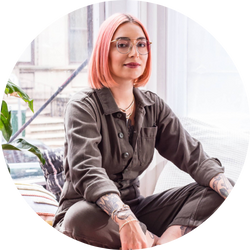 Angela Monaco, jewelry designer with pink hair, seated in a stylish pose in a casual outfit.
