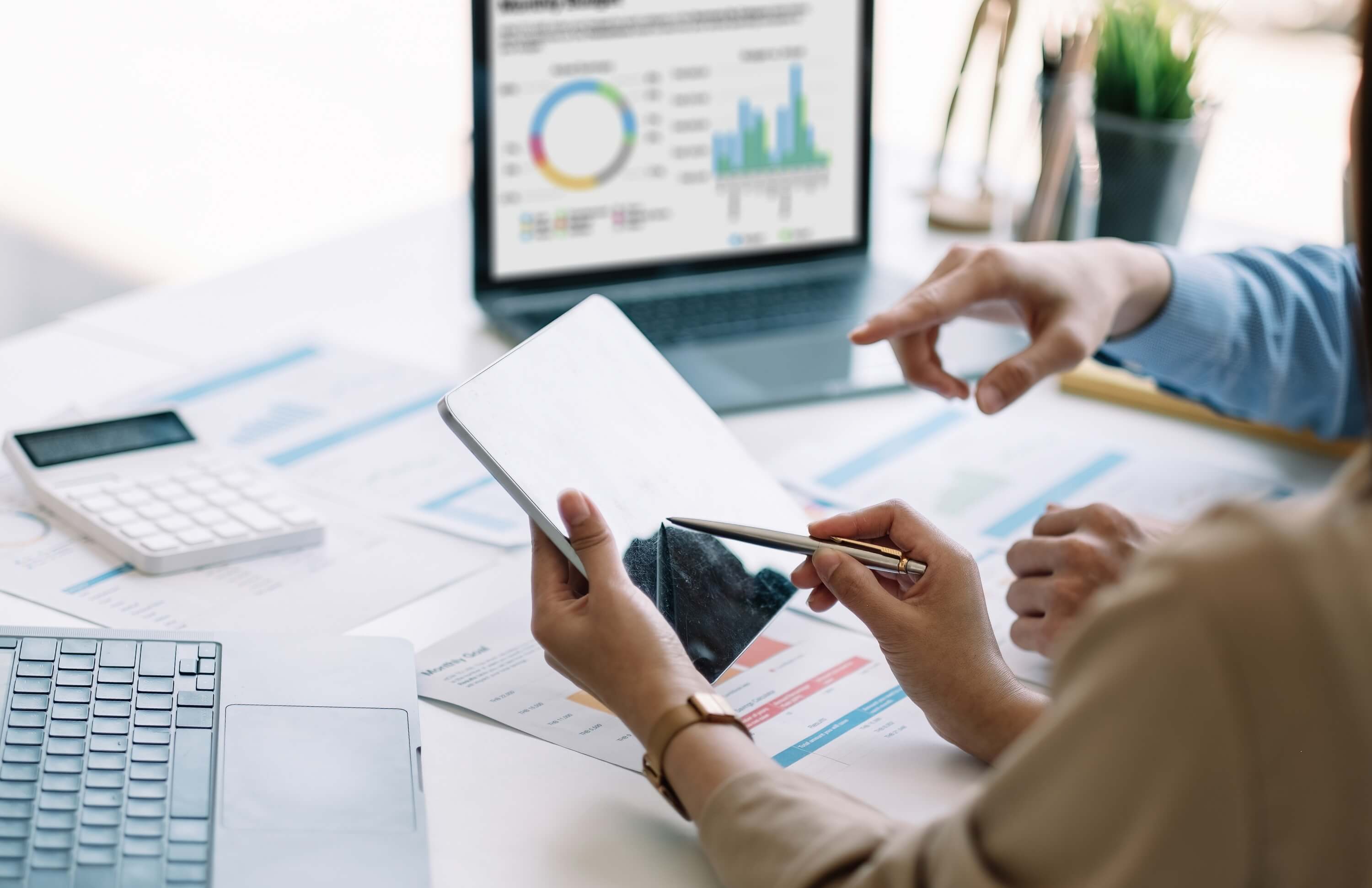 Hands using a tablet to review analytics data with charts and graphs on a laptop in the background.