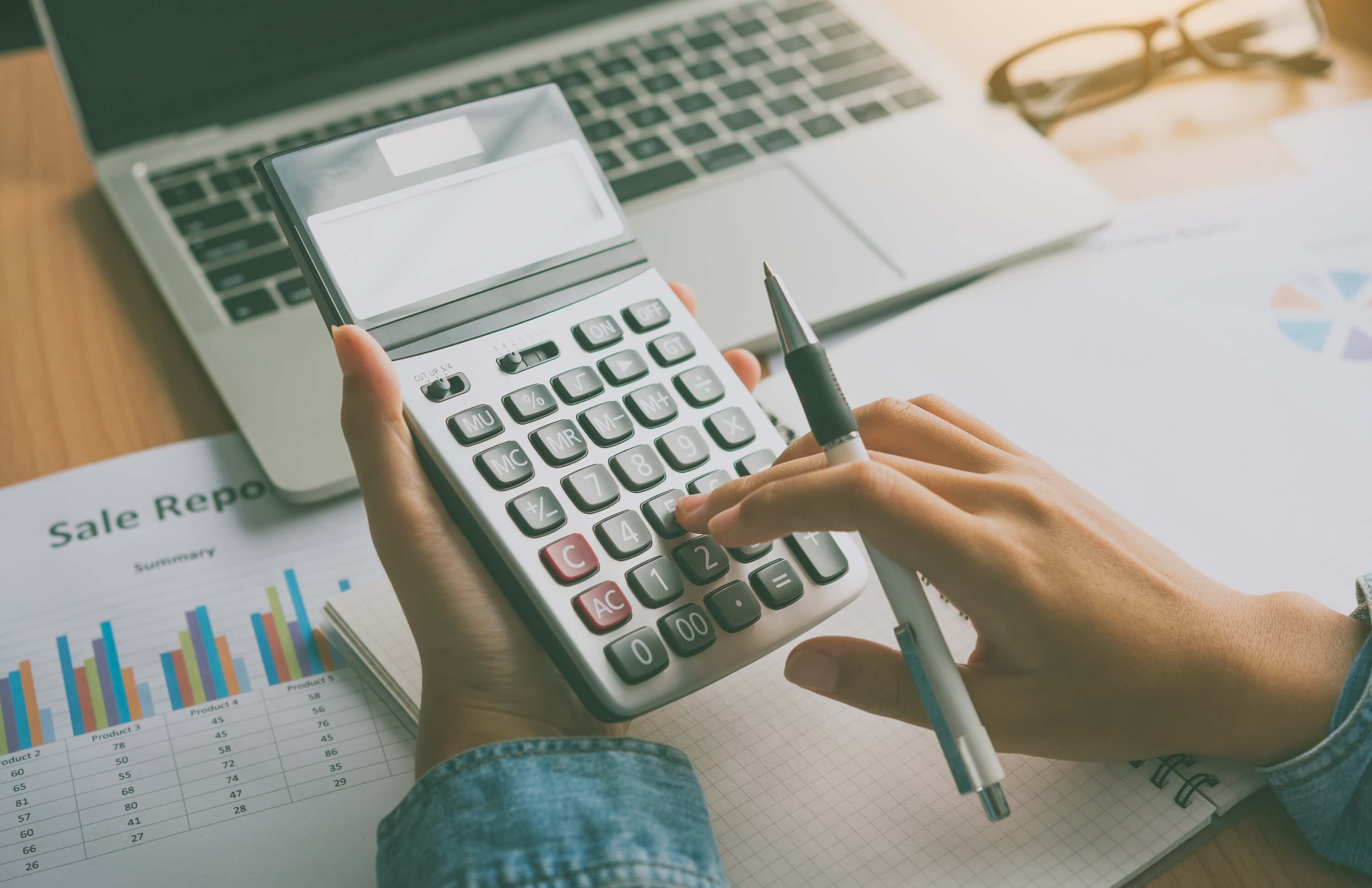 Hands using a calculator with sales reports and charts for dynamic pricing analysis.